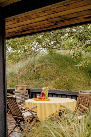 A wooden deck with a round table covered by a yellow tablecloth, surrounded by wooden chairs. - Nordic 70s gem amidst the dunes (Blokhus)