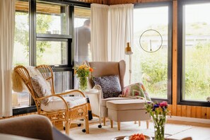 Ein Zimmer mit großem Fenster, einem Korbstuhl, einem kleinen Tisch mit Vase und einer Lampe.