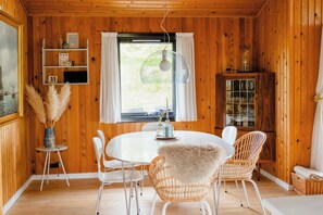 Ein Holzblockhaus-Interieur mit einem runden Esstisch, weißen Stühlen, einem Korbstuhl mit Felldecke, einem kleinen Beistelltisch mit einer Vase und einem Schrank mit Glastüren.