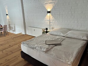 Un dormitorio con pared de ladrillo blanco, suelo de madera, cama con ropa de cama blanca, mesita de noche con lámpara y una silla.
