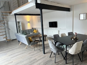 A modern living room with a grey sofa, a dining table with chairs, a staircase, and a TV mounted on the wall. - Stylish apartment in Baesweiler - near Aachen & Euregio from 129€ for 3 people (Baesweiler)