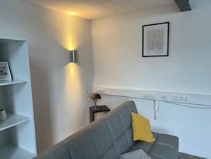 A room with a grey sofa, a yellow pillow, a framed picture, a wall-mounted light, and a power outlet. - Stylish apartment in Baesweiler - near Aachen & Euregio from 129€ for 3 people (Baesweiler)