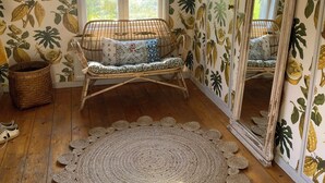 Una habitación con piso de madera, alfombra tejida, silla de mimbre con cojines, un espejo grande y una ventana con vista a follaje.