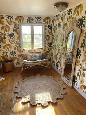 Ein Zimmer mit Holzboden, einem gewebten Teppich, einem Korbstuhl mit Kissen, einem großen Spiegel und einem Fenster mit Blick ins Grüne.