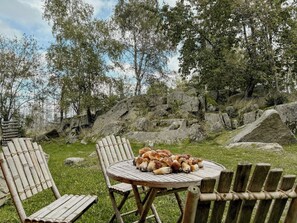 Ein runder Holztisch mit verschiedenen Backwaren, umgeben von Holzstühlen in einer grünen Umgebung im Freien mit Bäumen und Steinen im Hintergrund.