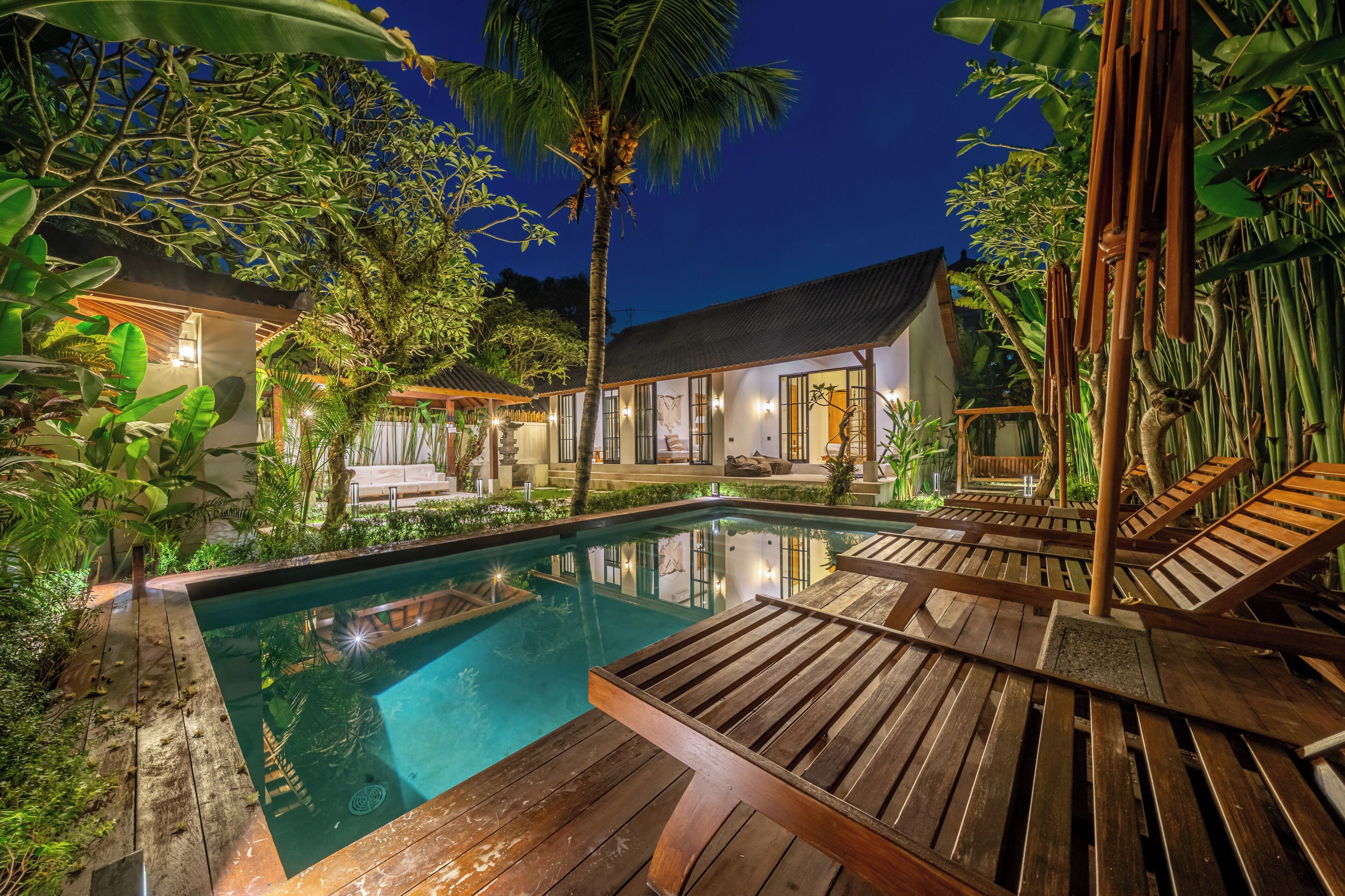 A swimming pool with wooden deck loungers, surrounded by lush greenery and a house with large windows.