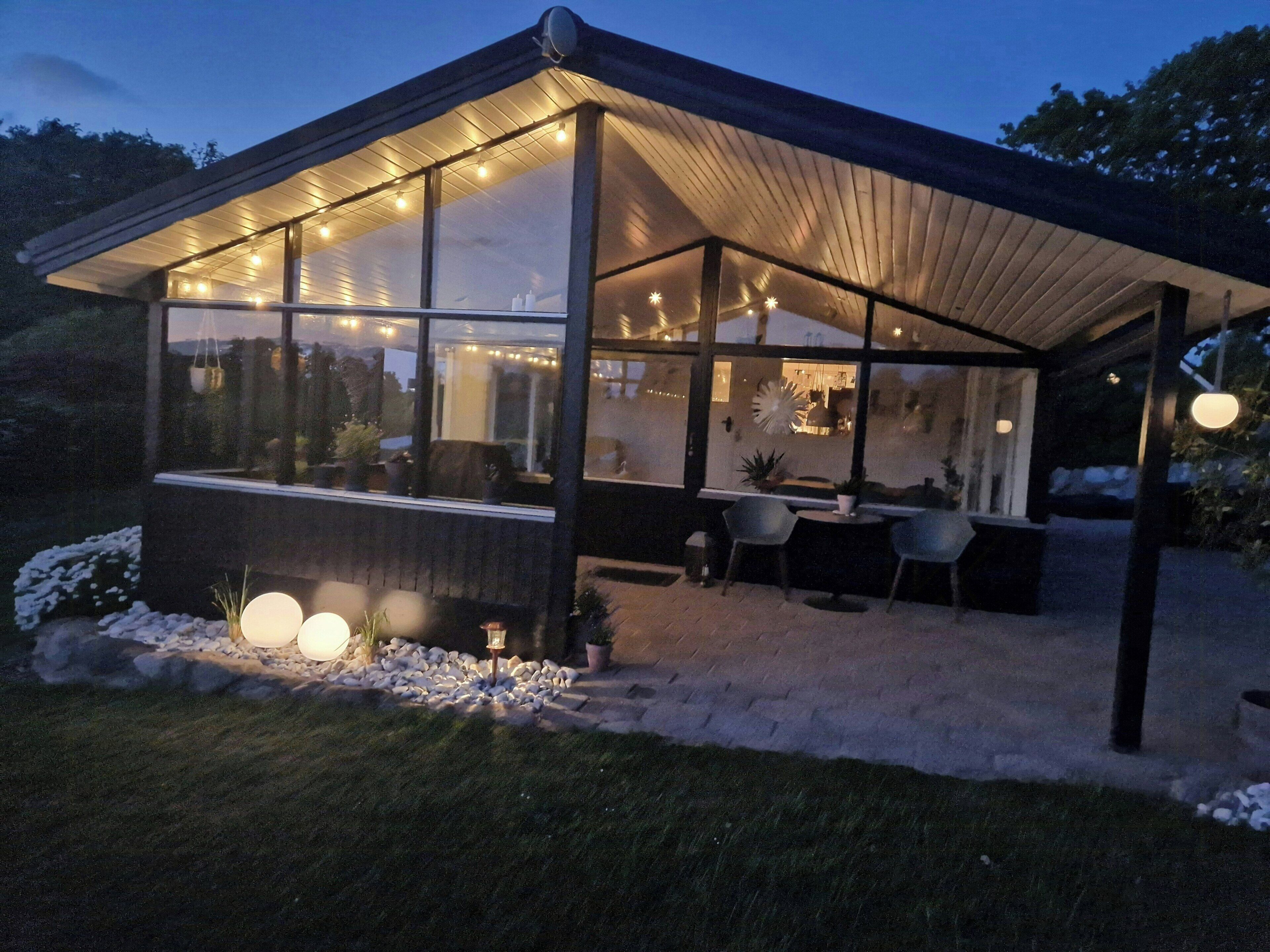 Una estructura moderna al aire libre con paredes de vidrio, una zona de estar techada y luces colgantes.