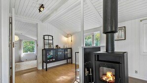 Une pièce avec un poêle à bois, des murs lambrés peints en blanc, une vitrine et une vue sur la verdure à l’extérieur.
