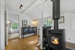 Ein Zimmer mit Kaminofen, weißen Wandverkleidungen, einer Glasschrank und Blick auf Grün.
