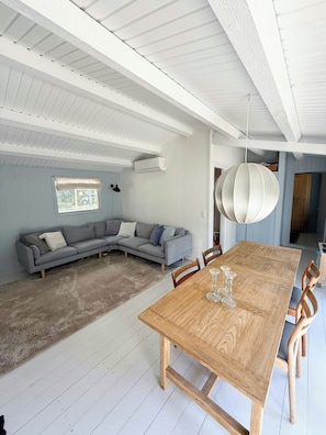 A modern living room with a wooden dining table, a grey sectional sofa, and a hanging light fixture. - Wooden cottage near the beach in Odsherred (Asnæs)