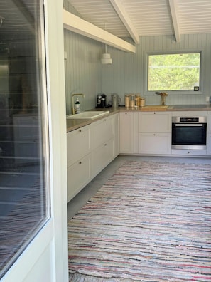 A kitchen with white cabinets, a built-in oven, and a window with a view of trees. - Wooden cottage near the beach in Odsherred (Asnæs)