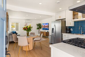 Eine moderne Küche mit einem Edelstahlkühlschrank, einem eingebauten Backofen und einem Gasherd. Es gibt einen Essbereich mit einem runden Tisch und Stühlen, und eine Couch im Hintergrund.