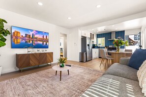 Ein modernes Wohnzimmer mit Flachbildfernseher, einer Couch, einem Couchtisch und einer Küche im Hintergrund.