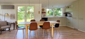 Ein Essbereich mit Holztisch und Stühlen, eine Küche mit Schränken und Geräten sowie Blick ins Grüne.