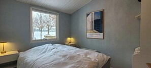 Ein Schlafzimmer mit Bett, einem Fenster mit Blick auf Bäume und Wasser, einer Lampe und einem gerahmten Bild an der Wand.