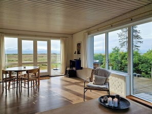 A room with a wooden floor, a round table with chairs, a wicker chair with cushions, a large window with a view of the countryside, and a small table with candles. - Beach house with sea view near Hou beach (Hals)
