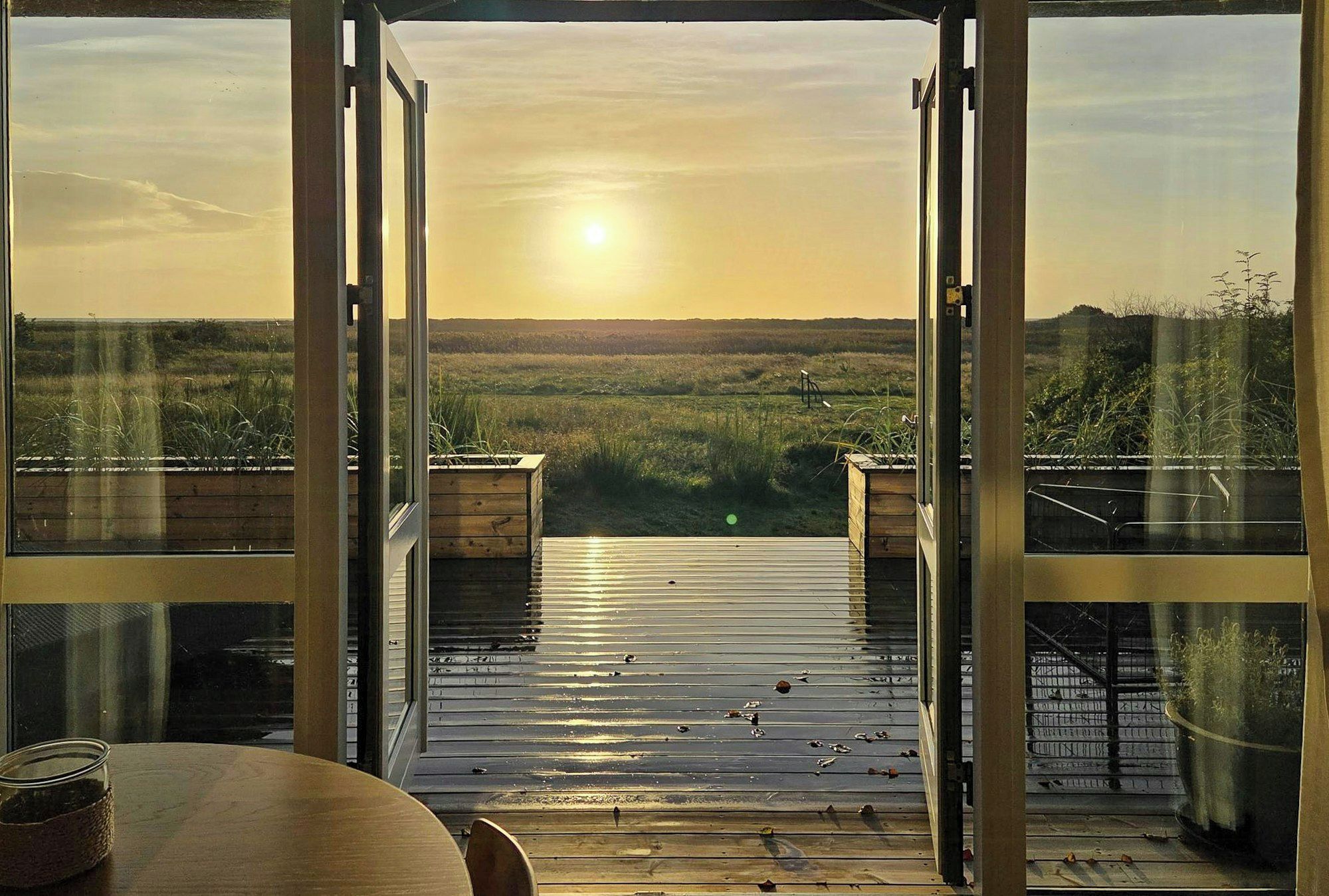 Blick von einem Zimmer mit großen Glasfenstertüren auf eine Terrasse im Freien, mit Ausblick auf eine grüne Landschaft bei Sonnenuntergang.