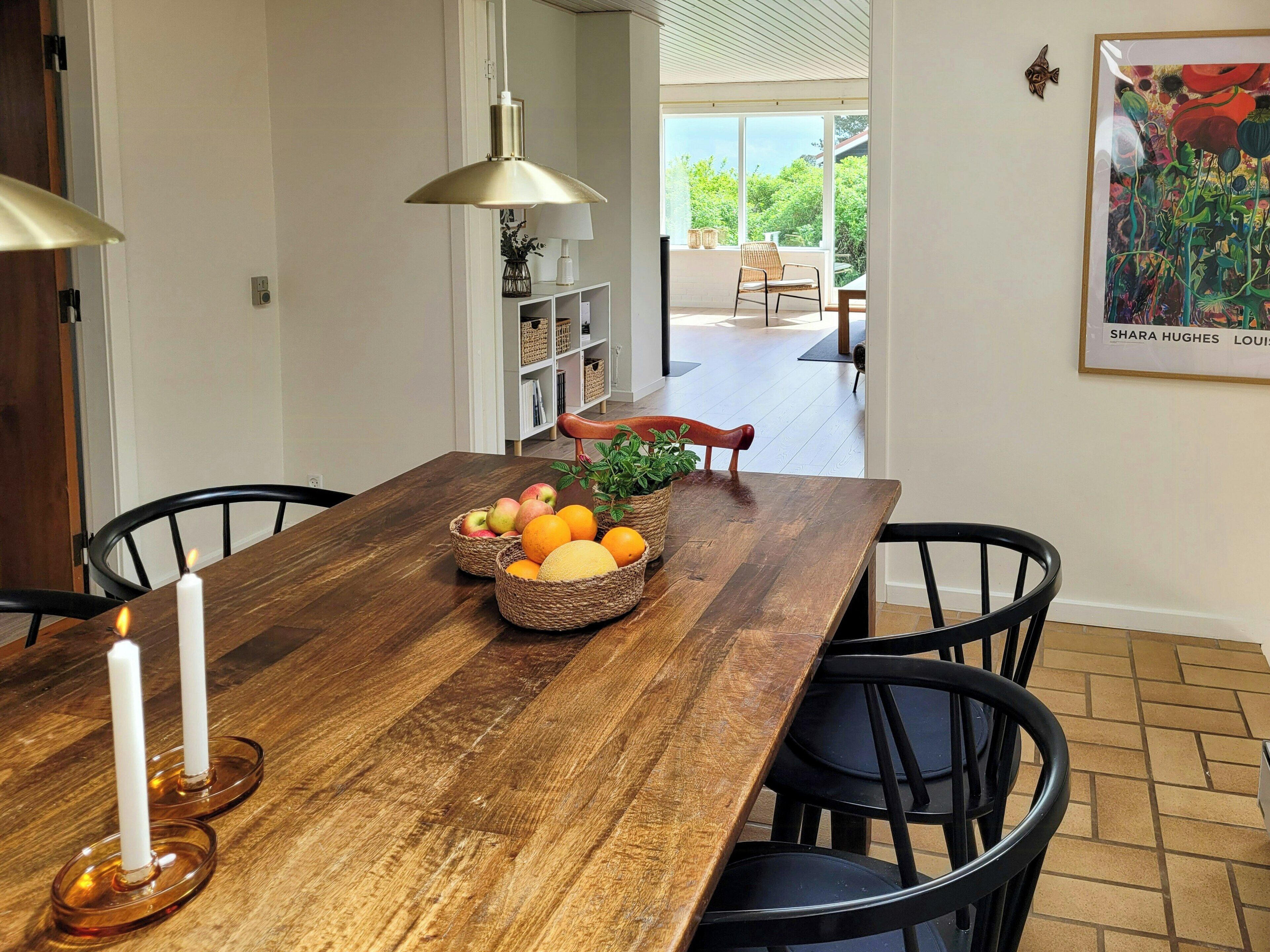 Een eetgedeelte met een houten tafel, zwarte stoelen, een kaars, fruitmanden en een hanglamp.