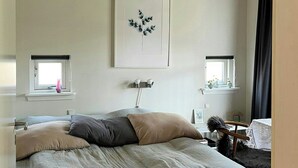 Un dormitorio con cama, ventana con vistas, un cuadro en la pared y una mesita de noche.