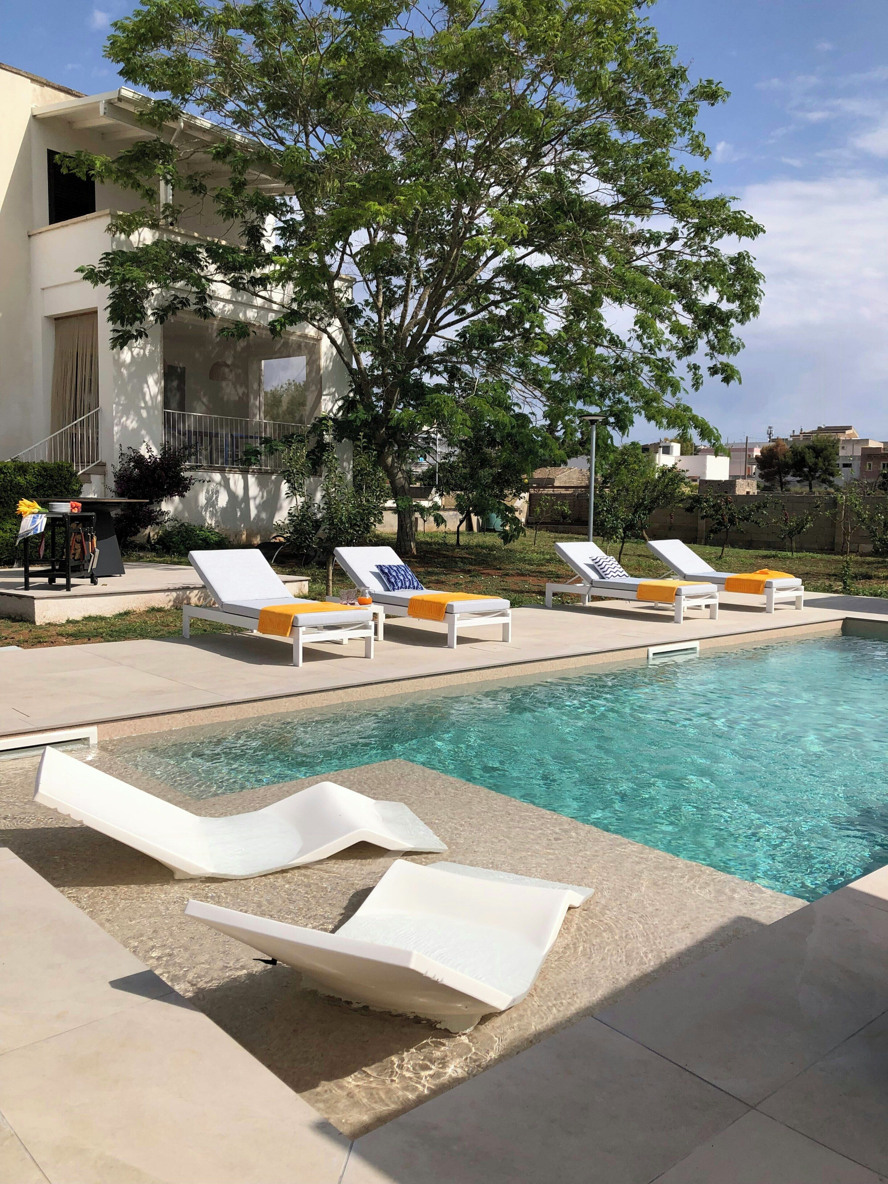 Un área de piscina con sillas de descanso, un árbol y un edificio al fondo.