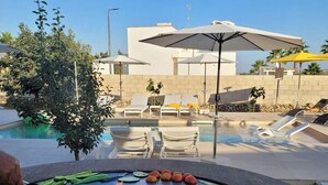 Ein Grill am Pool mit Gemüse, Feuer, Liegestühlen, Sonnenschirmen und einem Gebäude im Hintergrund.