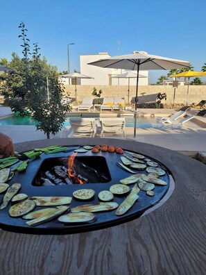 Ein Grill am Pool mit Gemüse, Feuer, Liegestühlen, Sonnenschirmen und einem Gebäude im Hintergrund.