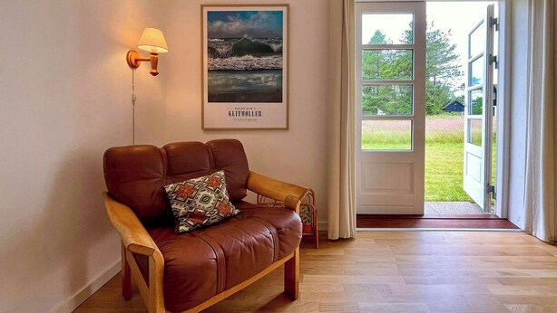 Una habitación con un sofá de cuero marrón, una lámpara de pared, un cuadro enmarcado con una escena costera y una puerta que da a un espacio al aire libre.
