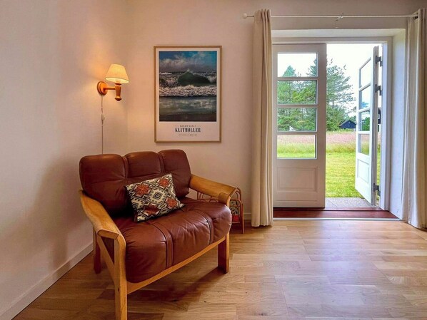 A room with a brown leather sofa, a wall-mounted lamp, a framed picture of a coastal scene, and a door leading to an outdoor area. - Retro holiday home by Thy National Park (Bedsted)