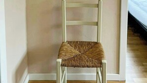 Una silla blanca de tejido en una habitación con suelo de madera, paredes color beige y una ventana que da a un balcón.