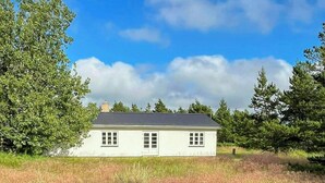 Una casa de un piso con fachada blanca y techo oscuro, rodeada por un campo cubierto de césped y árboles.