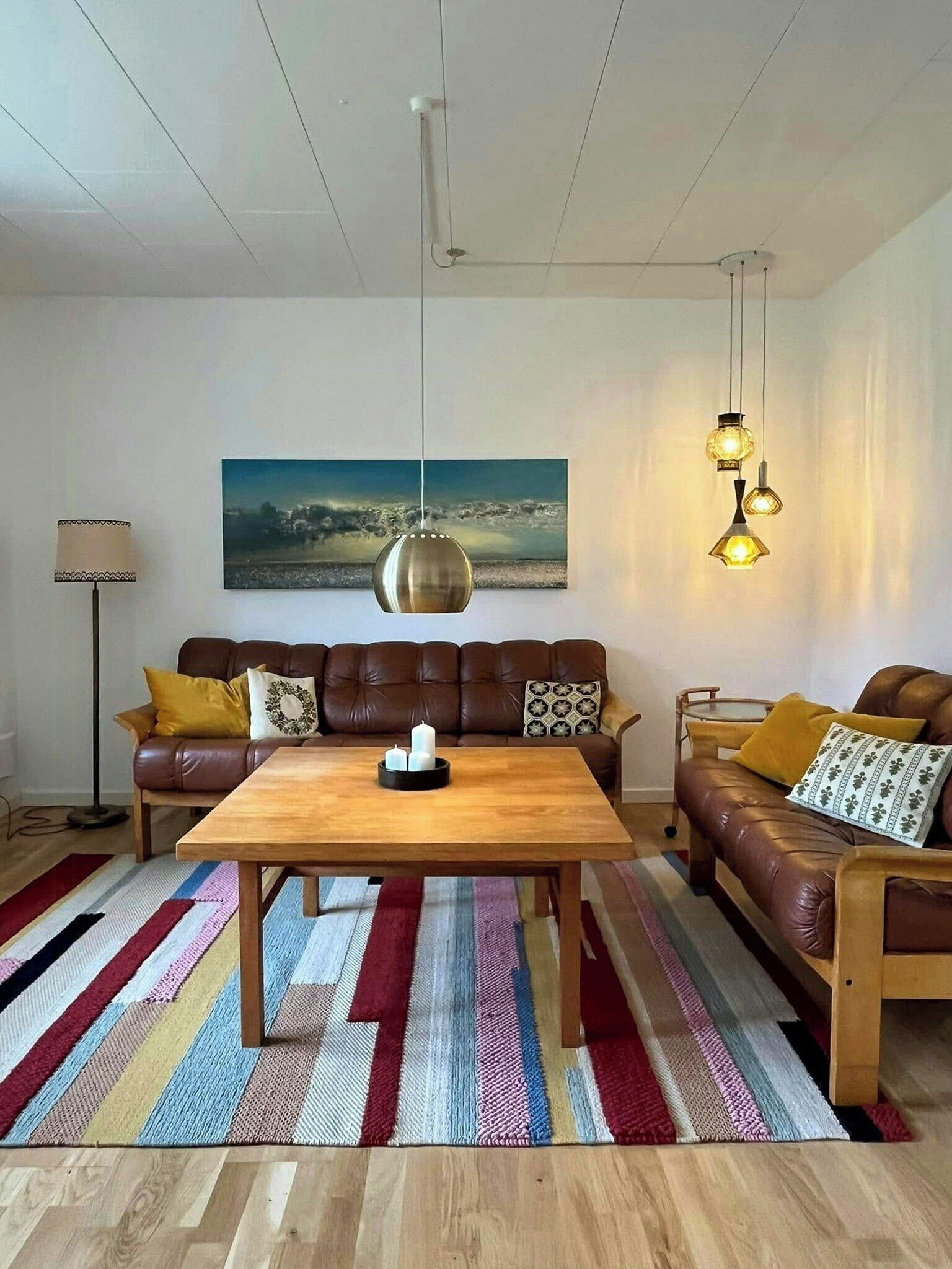 Un salon moderne avec une table basse en bois, des canapés en cuir, un tapis rayé aux couleurs vives et des luminaires suspendus.