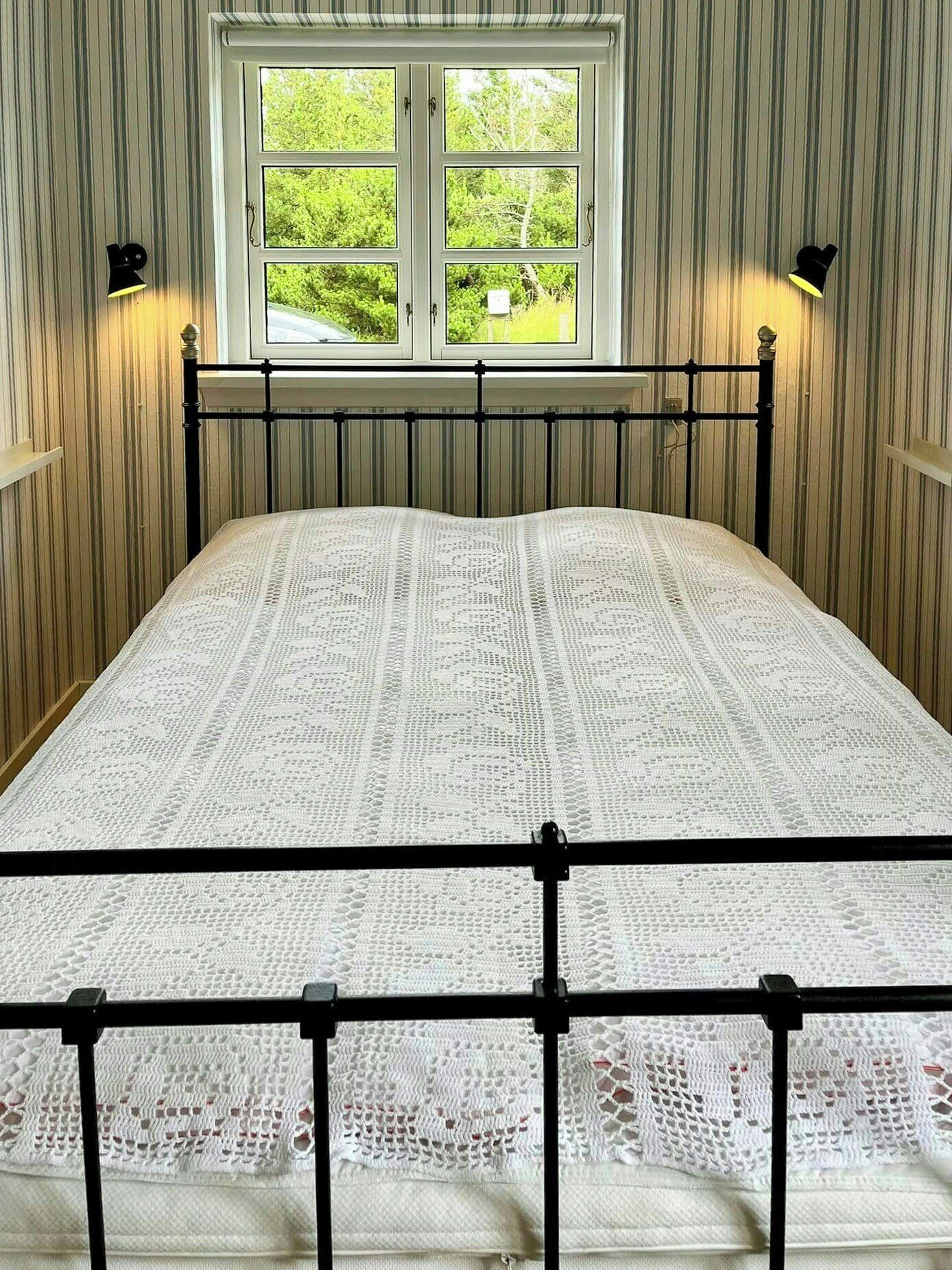 Ein Bett mit weißem Spitzenüberwurf, ein Fenster mit Blick auf Grünflächen und zwei Wandlampen.