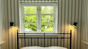 Una cama con una colcha de encaje blanco, una ventana con vistas a follaje, y dos lámparas fijadas a la pared.