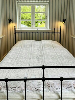 A bed with a white lace bedspread, a window with a view of greenery, and two wall-mounted lamps. - Retro holiday home by Thy National Park (Bedsted)