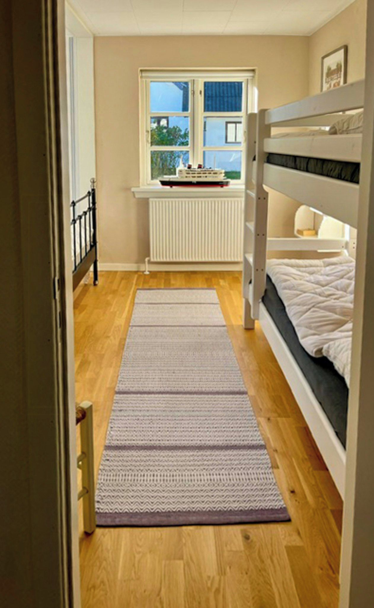 Un pasillo estrecho con piso de madera, una ventana que da a zonas verdes y una cama litera con ropa blanca.