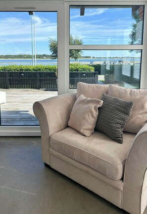 Un sofá beige con un cojín con textura en una habitación con ventanas amplias que dan a la costa.