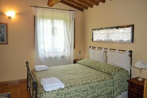 A bedroom with a bed, a nightstand, a lamp, a window with curtains, and a wall-mounted light fixture. - Private pool stone villa overlooking Crete Senesi (Asciano)