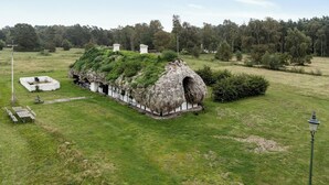 Une construction au toit de chaume, surmontée d’un toit végétalisé, entourée d’une pelouse bien entretenue et de quelques arbres en arrière-plan.