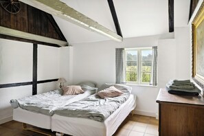 A bedroom with a single bed, a wooden dresser, a window with a view, and exposed wooden beams. - Renovated stable with seaweed roof on Læsø (Læsø)