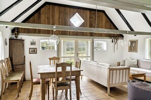A rustic interior with a wooden dining table, chairs, a sofa, and a large window. - Renovated stable with seaweed roof on Læsø (Læsø)