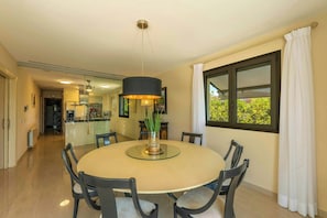 Un espace repas moderne avec une table ronde en verre, six chaises, un luminaire central et une vue dégagée sur l’extérieur grâce à une grande fenêtre.