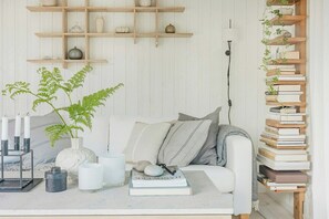 Ein gemütliches Wohnzimmer mit einem weißen Sofa, einem Couchtisch mit Büchern und Kerzen, einer Pflanze und einem Holzregal.