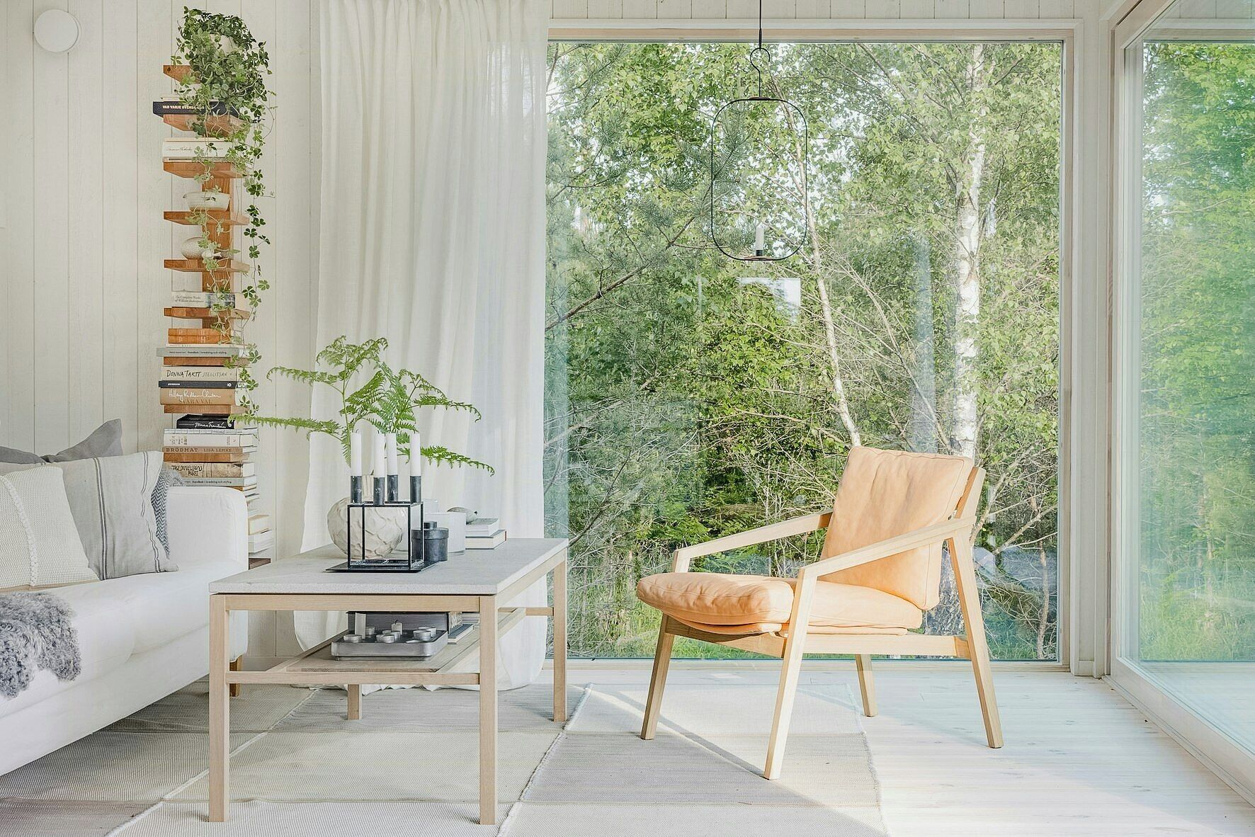 A modern living room with a sofa, a table with a vase and candles, a chair, and a view of trees through large windows.