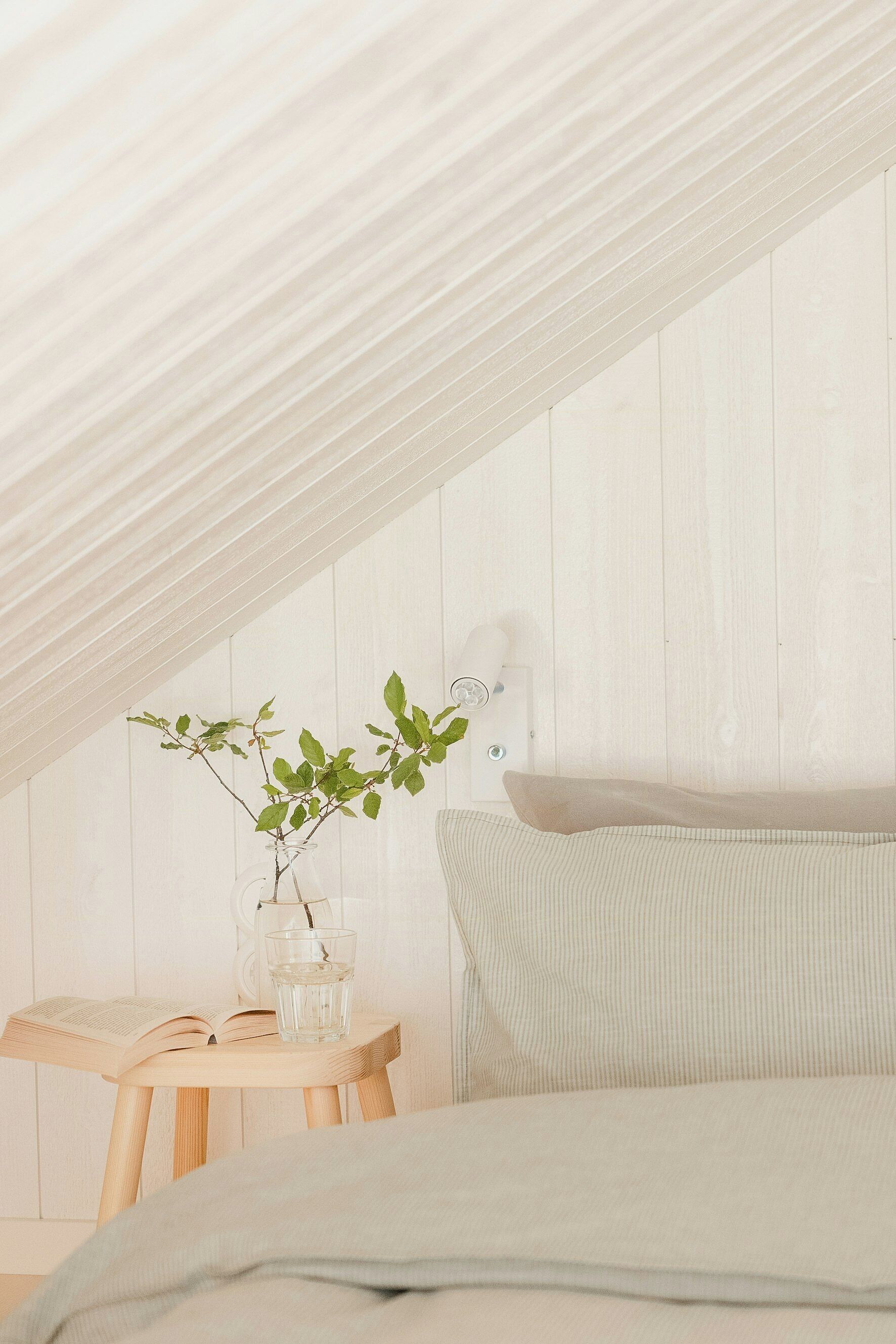 A bedroom with a sloped ceiling, a bed, a bedside table with a glass of water, and a book.