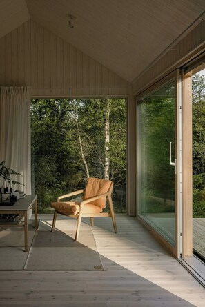 Ein modernes Interieur mit Holzboden, eine große Glasschiebetür zu einem bewaldeten Gebiet, ein Holzstuhl und ein kleiner Tisch mit einer Pflanze.