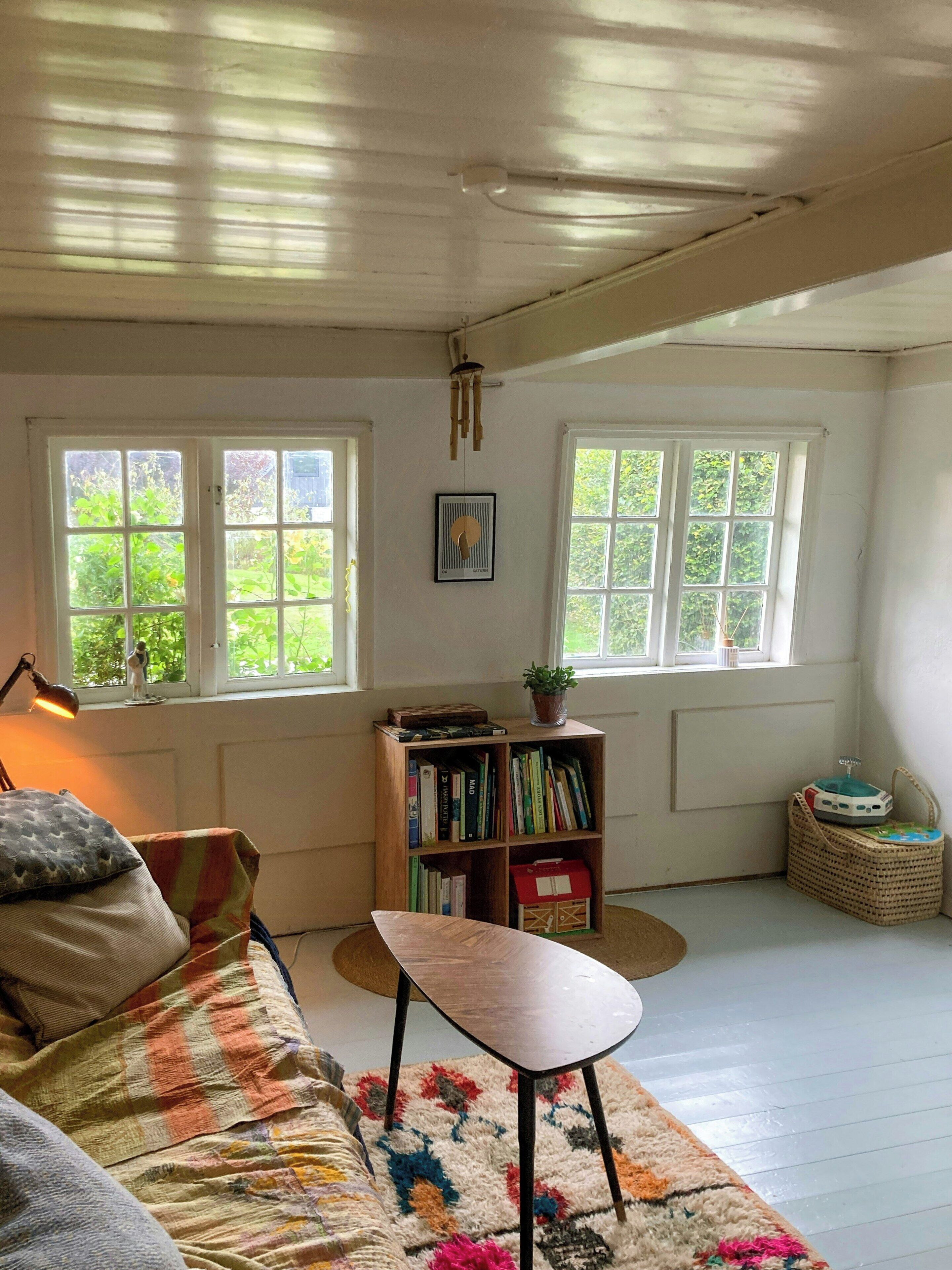 Een kamer met een houten vloer, een bank met een kleurrijk kleed, een boekenkast en twee ramen met uitzicht op groen.