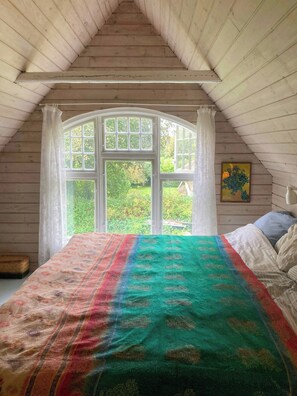 Ein Schlafzimmer mit großem Fenster, einem Bett mit bunter Decke und einer Holzzieldecke.