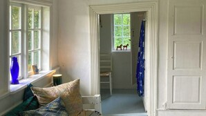 Une pièce avec un plancher en bois blanc, une porte blanche portant l’inscription « HABS », une fenêtre donnant sur un paysage verdoyant et un lit agrémenté d’oreillers à motifs.