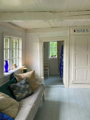 Ein Zimmer mit Holzboden in Weiß, einer weißen Tür mit der Aufschrift 'HABS', einem Fenster mit Blick auf Grünflächen und einem Bett mit gemusterten Kissen.