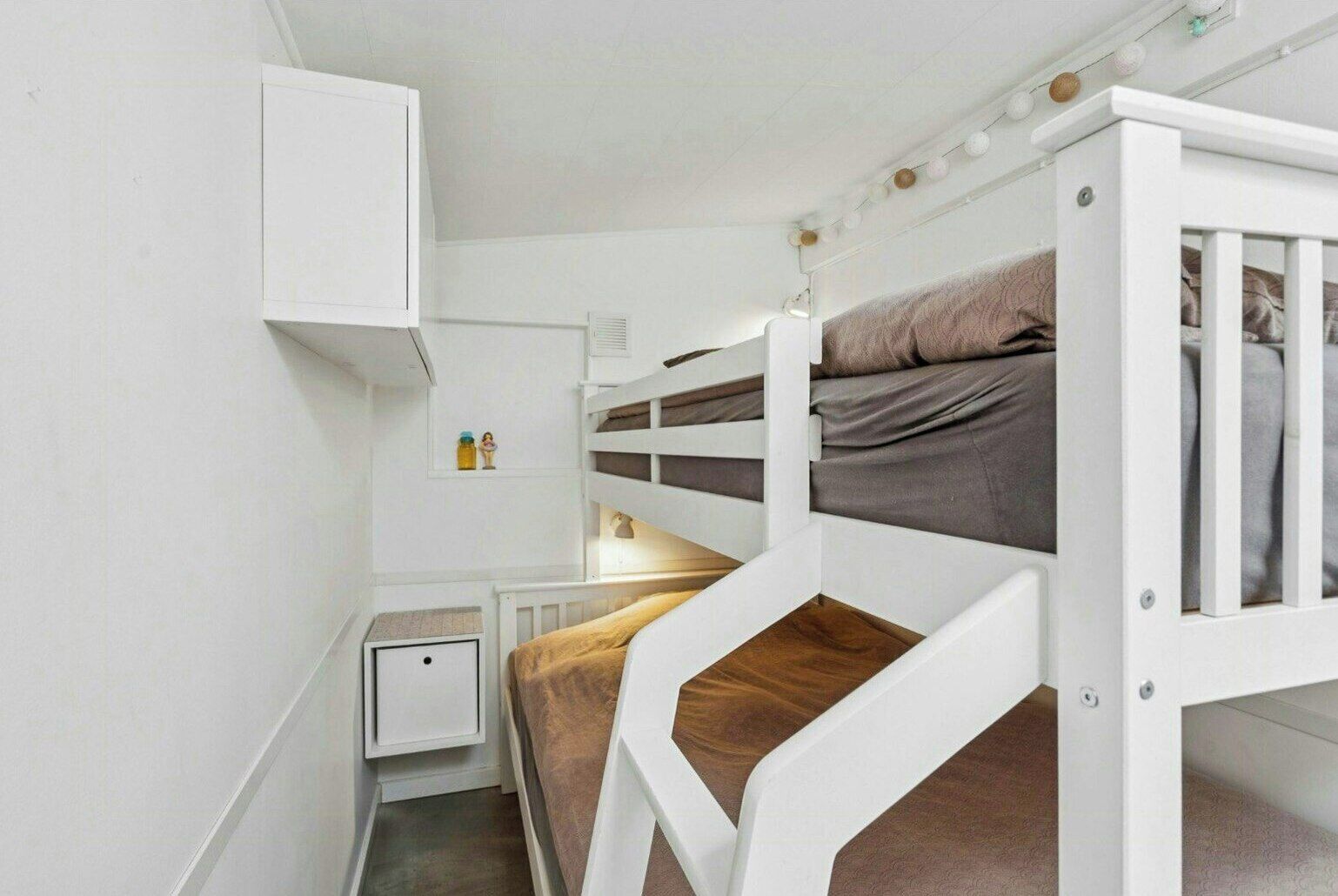 A bunk bed room with a white wardrobe, bedside table, and a small shelf with items.